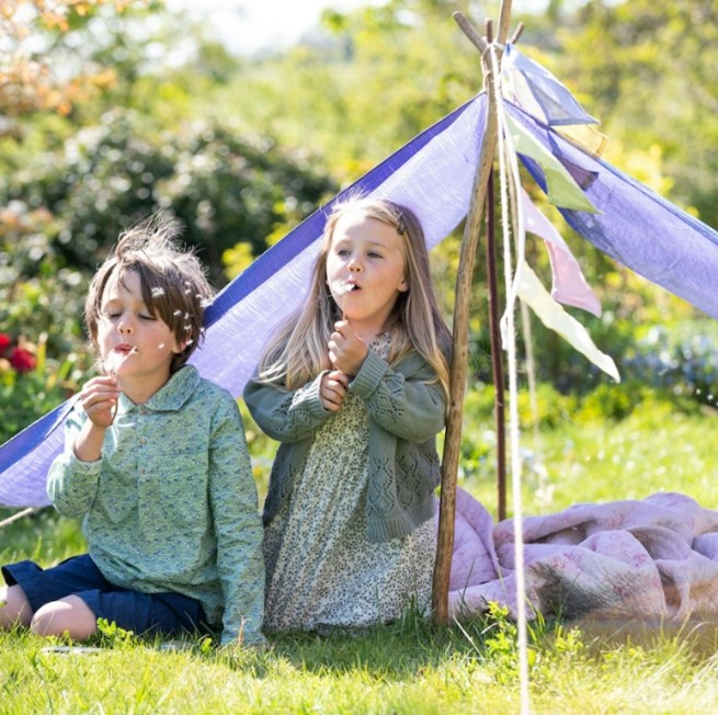 Young girl and boy blowing dandelions outside but under a purple tent